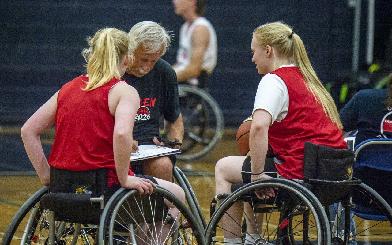 A photo of two wheelchair basketball athletes getting instruction from a coach on a basketball court